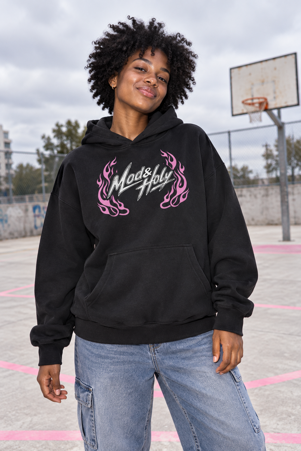 Young adult woman wearing the pink-flame Mod and Holy hoodie at a basketball court.